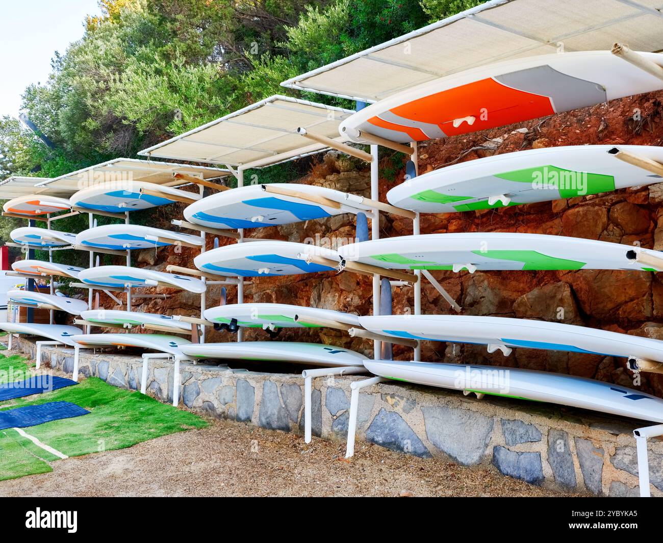 Molte tavole da surf colorate sono impilate in modo ordinato su una griglia metallica sotto un baldacchino, pronte per l'uso in acqua da parte dei windsurf Foto Stock