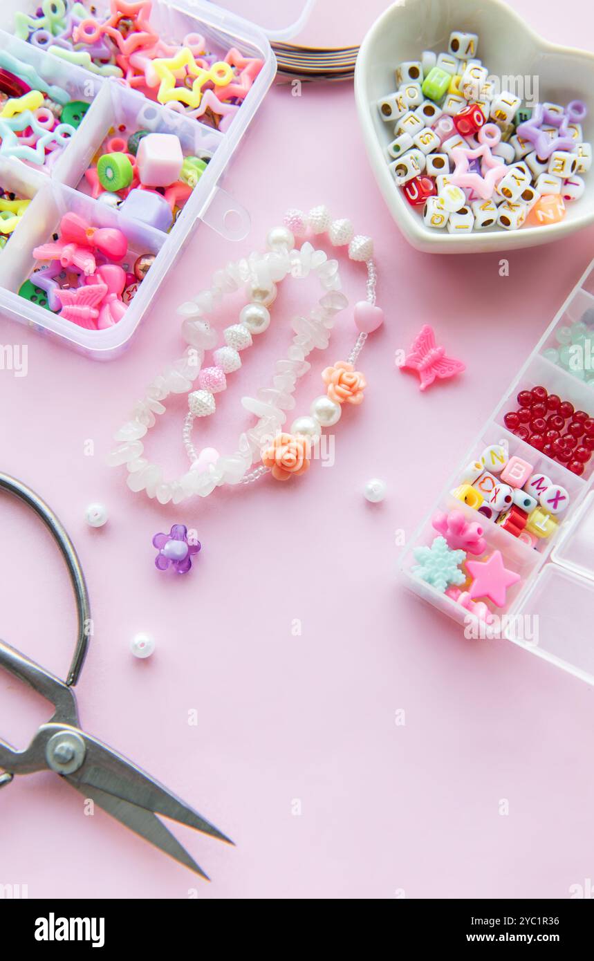 Una disposizione creativa di perline colorate, ciondoli e forbici su una superficie rosa pastello mostra il processo di creazione di gioielli unici. Flora delicata Foto Stock