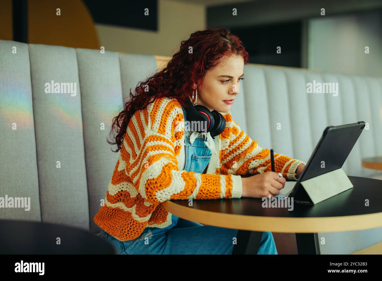Una studentessa studia con cura con un tablet, indossa cuffie e un maglione colorato a righe, seduto in un ambiente accogliente e moderno. Foto Stock