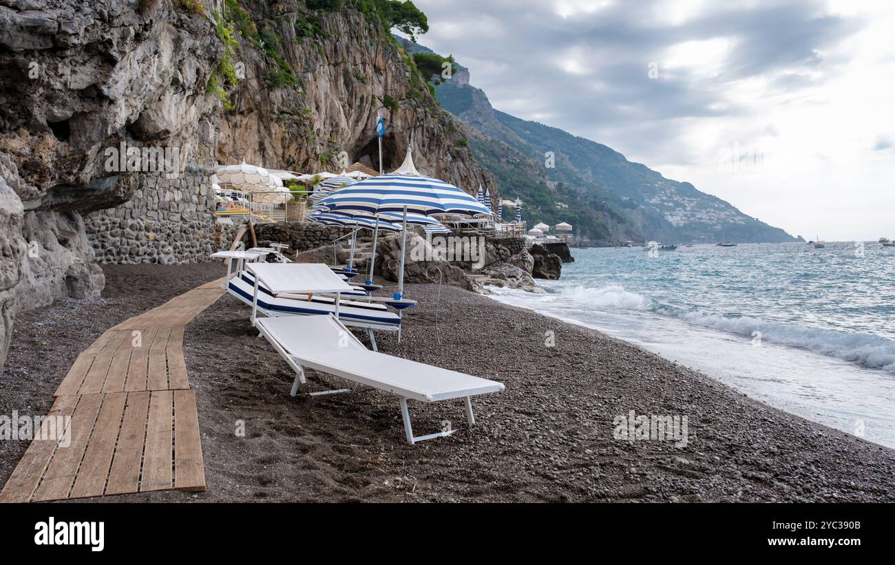 I visitatori si rilassano lungo la spiaggia ricoperta di ciottoli della Costiera Amalfitana, godendo di splendide viste, ombrelloni a righe e tranquille onde che si infrangono sulla riva Foto Stock