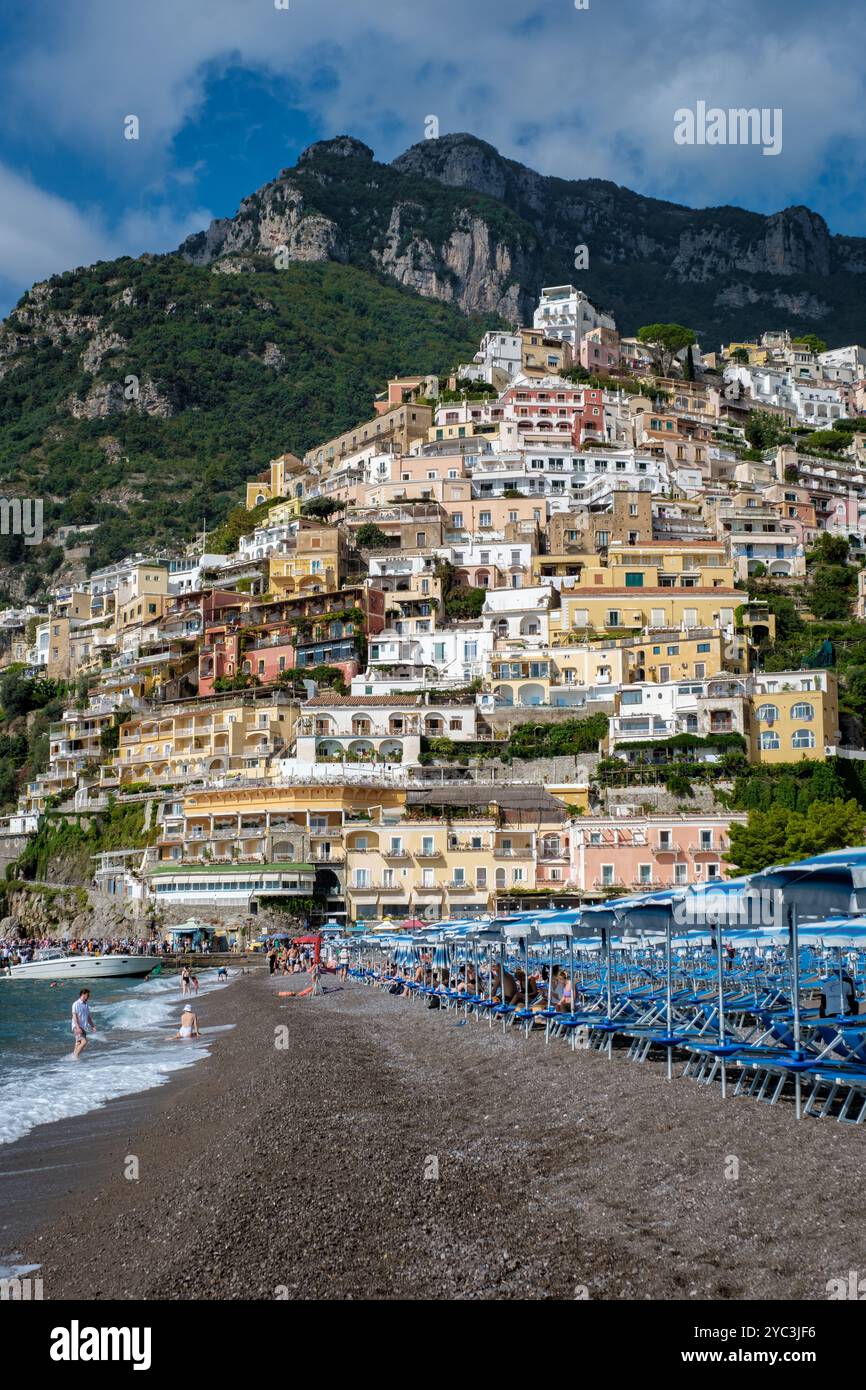 Le case colorate scendono lungo la collina di Positano, mentre le dolci onde si infrangono sulla spiaggia di ciottoli, offrendo una giornata perfetta sotto il sole di Amalfi. Foto Stock