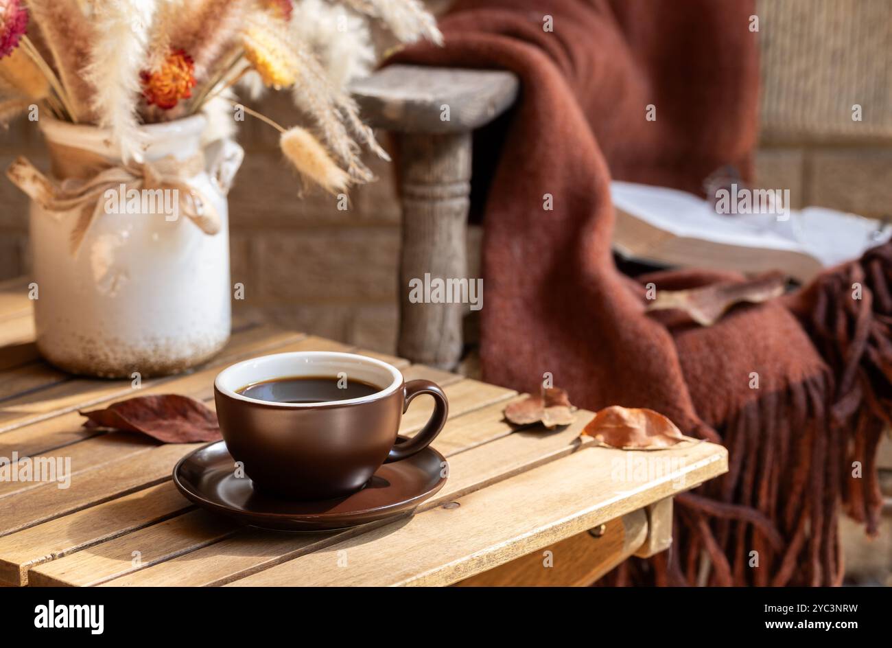 Tazza di caffè con vaso di fiori secchi e foglie autunnali su un tavolo di legno e una vecchia sedia con coperta sullo sfondo Foto Stock