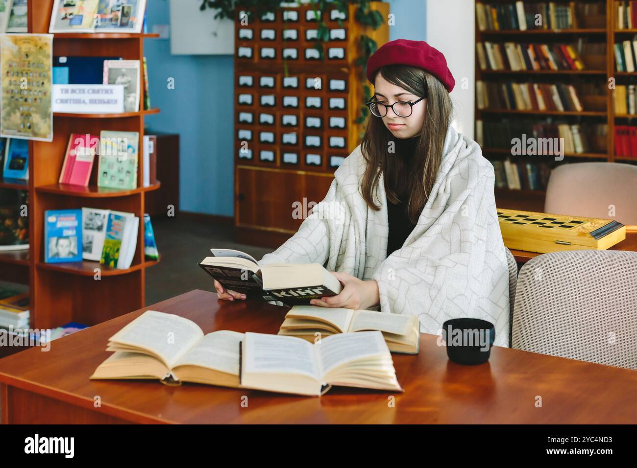 Romny, Ucraina, 29 settembre 2019: Una giovane donna siede in un ambiente accogliente della biblioteca e legge un libro, avvolto in una coperta, con un'espressione stanca su di lui Foto Stock