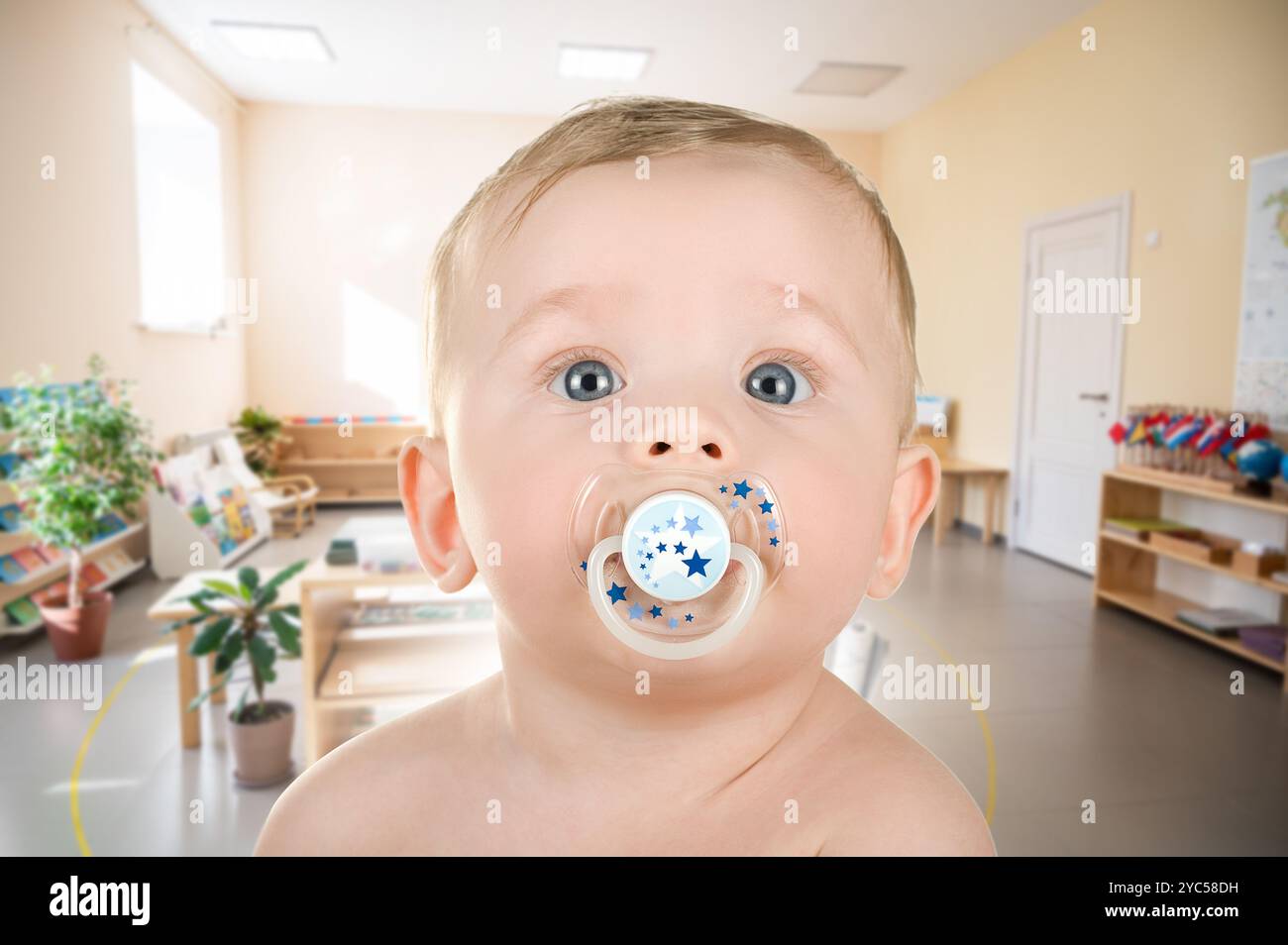 Primo piano di un bambino con un succhietto in un'aula luminosa o in un ambiente di cura diurna, circondato da materiale didattico. Foto Stock