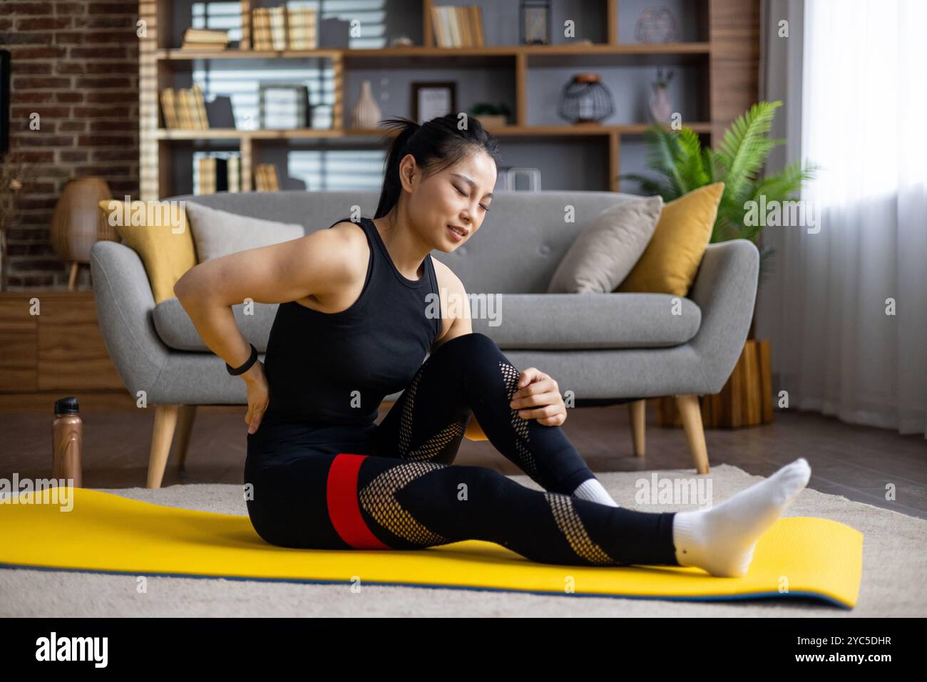 Donna asiatica che soffre mal di schiena seduta su un tappetino da yoga in salotto. Indossando abiti da allenamento neri, si allunga delicatamente per il sollievo. Concentratevi sulla salute AN Foto Stock