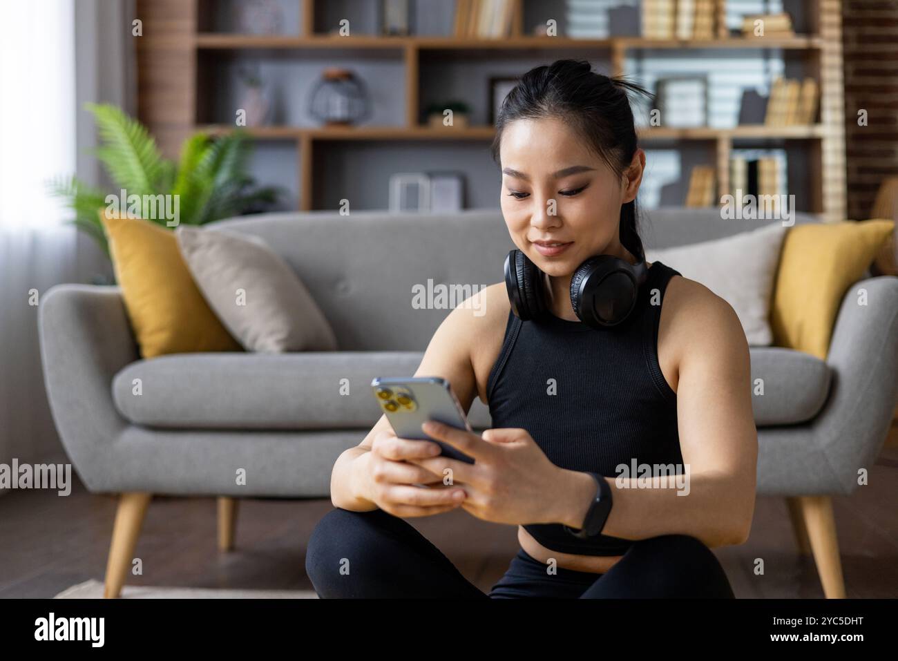 Donna asiatica in abbigliamento sportivo esegue esercizi, guarda corsi di fitness online sul telefono, cuffie intorno al collo, in un ambiente confortevole. Impegnato in guarigione Foto Stock