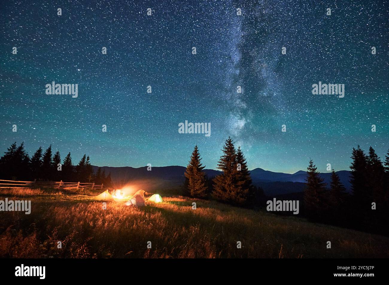 Sagome di turisti che riposano in campeggio intorno al fuoco con tende luccicanti nelle vicinanze sotto il cielo stellato. La via Lattea si estende in alto e getta una luce serena e magica sul tranquillo paesaggio montano. Foto Stock