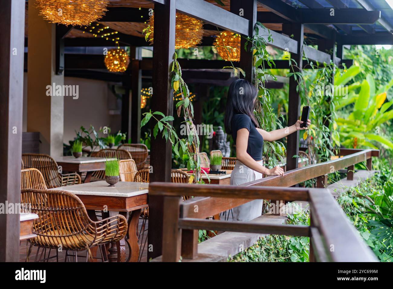 Una donna che si prende cura di sé in un ristorante tropicale. L'area esterna presenta mobili in legno, vegetazione lussureggiante e illuminazione decorativa. L'atmosfera Foto Stock