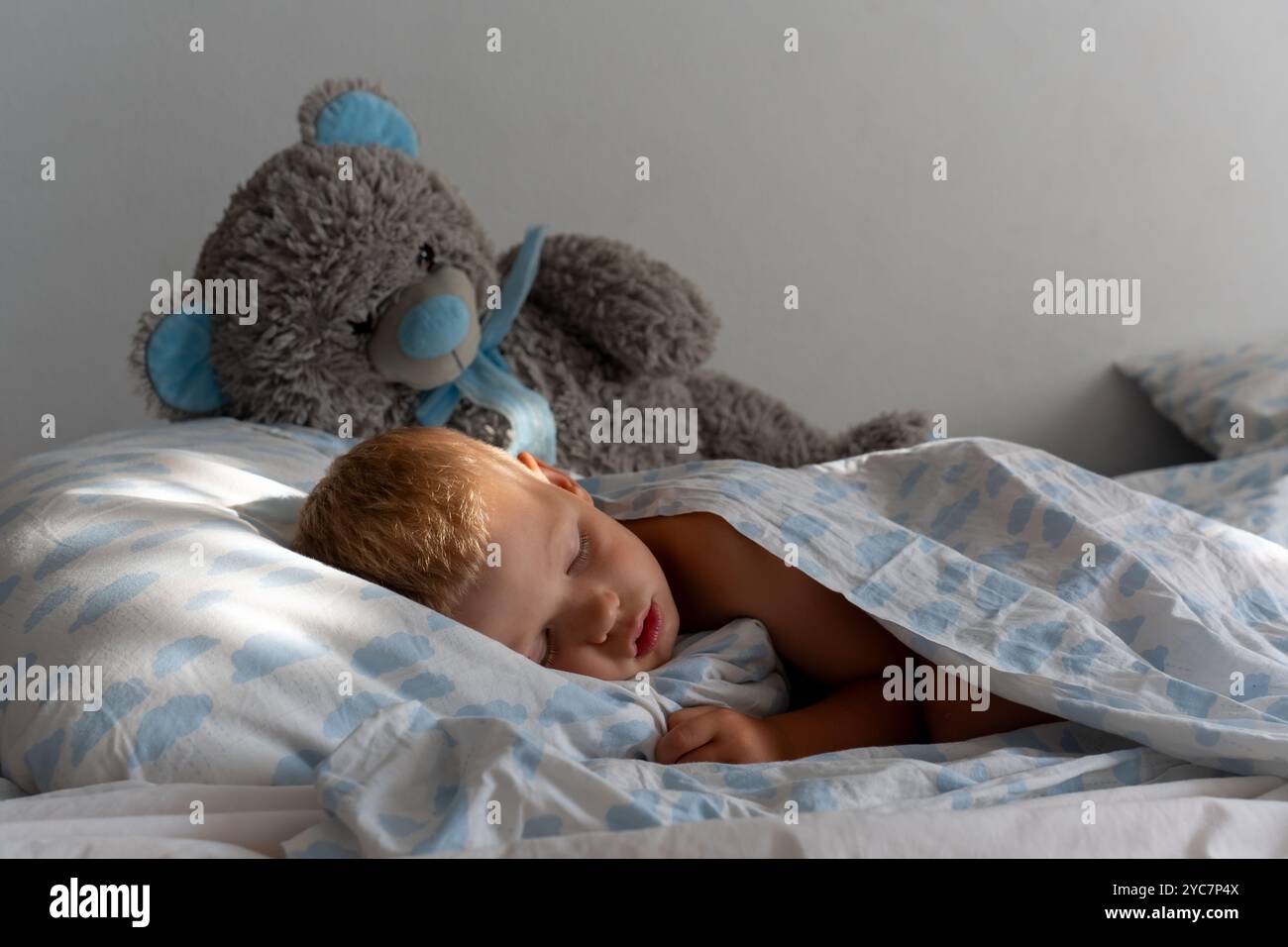 Un bambino simpatico suona addormentato nel letto con biancheria da letto blu a forma di nuvola e un grande orsacchiotto imbottito su una calma. Foto Stock