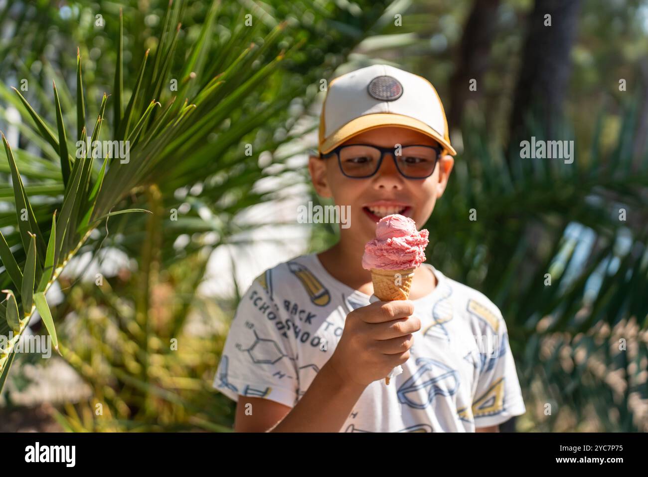 Ragazzo felice che indossa occhiali e un berretto, tenendo in mano un cono di gelato alle fragole in una brillante giornata estiva. Foto Stock