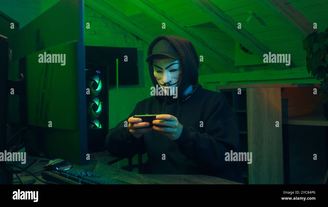 Leopoli, Ucraina - 3 giugno 2024: Uomo in maschera che usa il telefono cellulare smartphone anonimo guardando il programma di tecnologia della fotocamera furto sicuro pericolo bancario Foto Stock