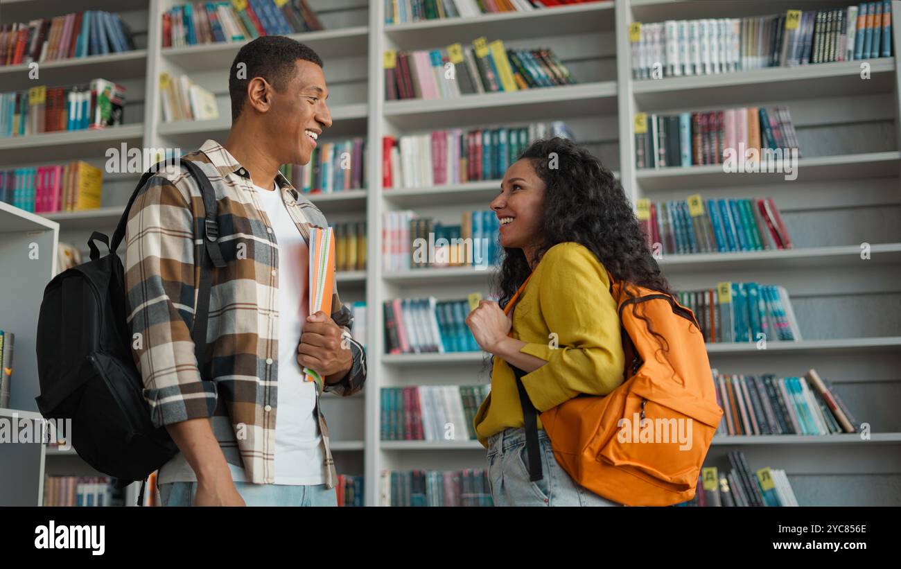 Due persone uomo afroamericano donna latino ispanica parlante biblioteca lettura club studenti di letteratura libreria scaffali di libreria comunicazione sorridente Foto Stock