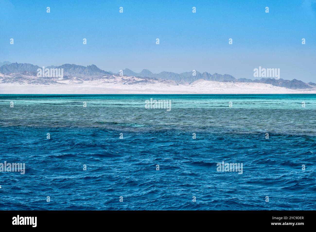 Vista mozzafiato del Mar Rosso con acque blu vibranti, che passano in sfumature turchesi poco profonde. Montagne aride e aspre sullo sfondo sotto il cielo limpido Foto Stock