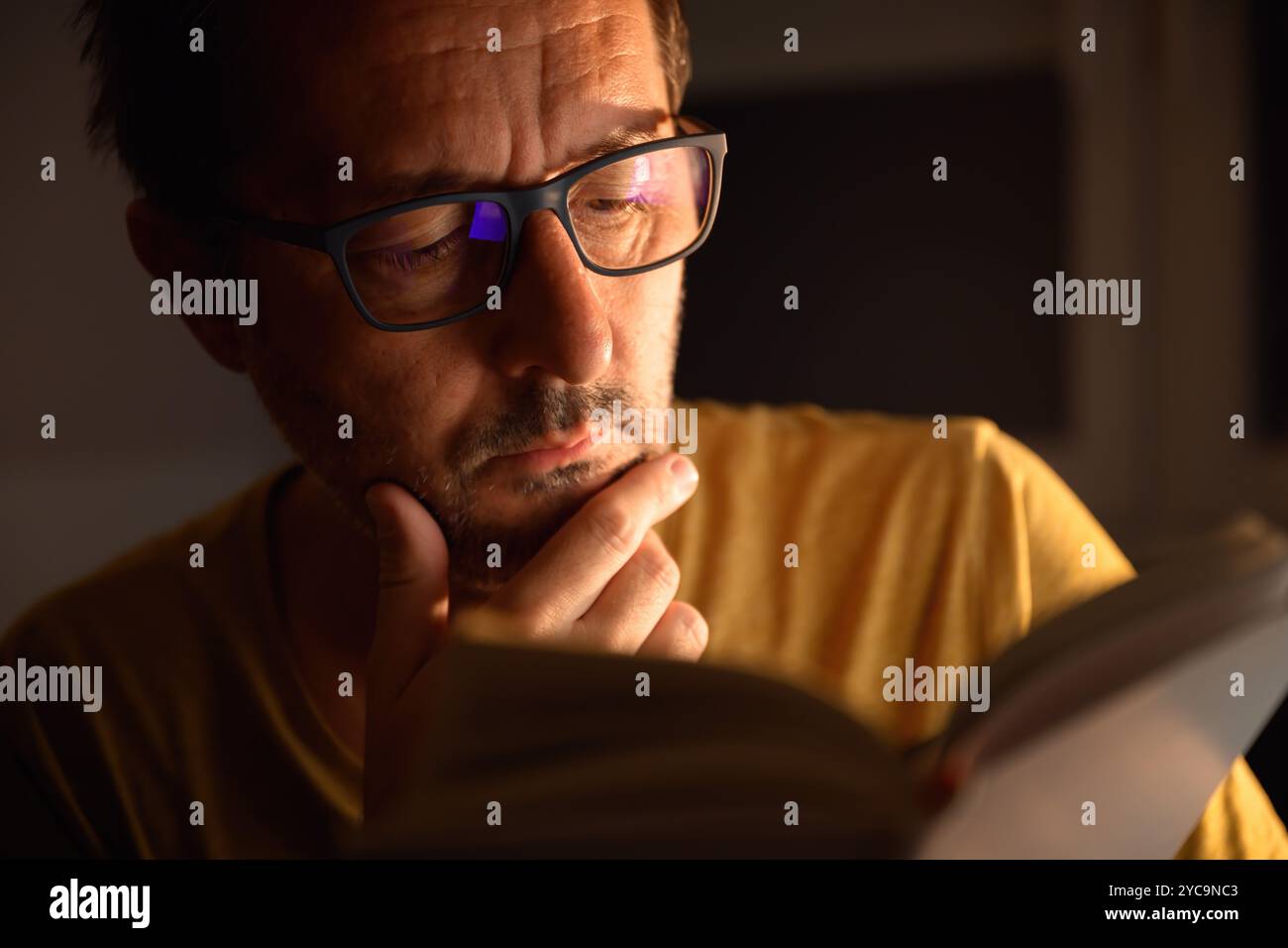 Bookworm che legge libri di letteratura di notte in camera buia, il suo volto è illuminato da una lampada da scrivania. Messa a fuoco selettiva. Foto Stock