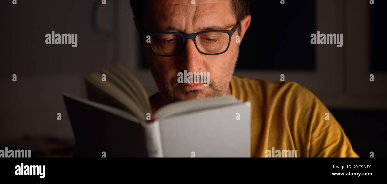 Bookworm che legge libri di letteratura di notte in camera buia, il suo volto è illuminato da una lampada da scrivania. Messa a fuoco selettiva. Foto Stock