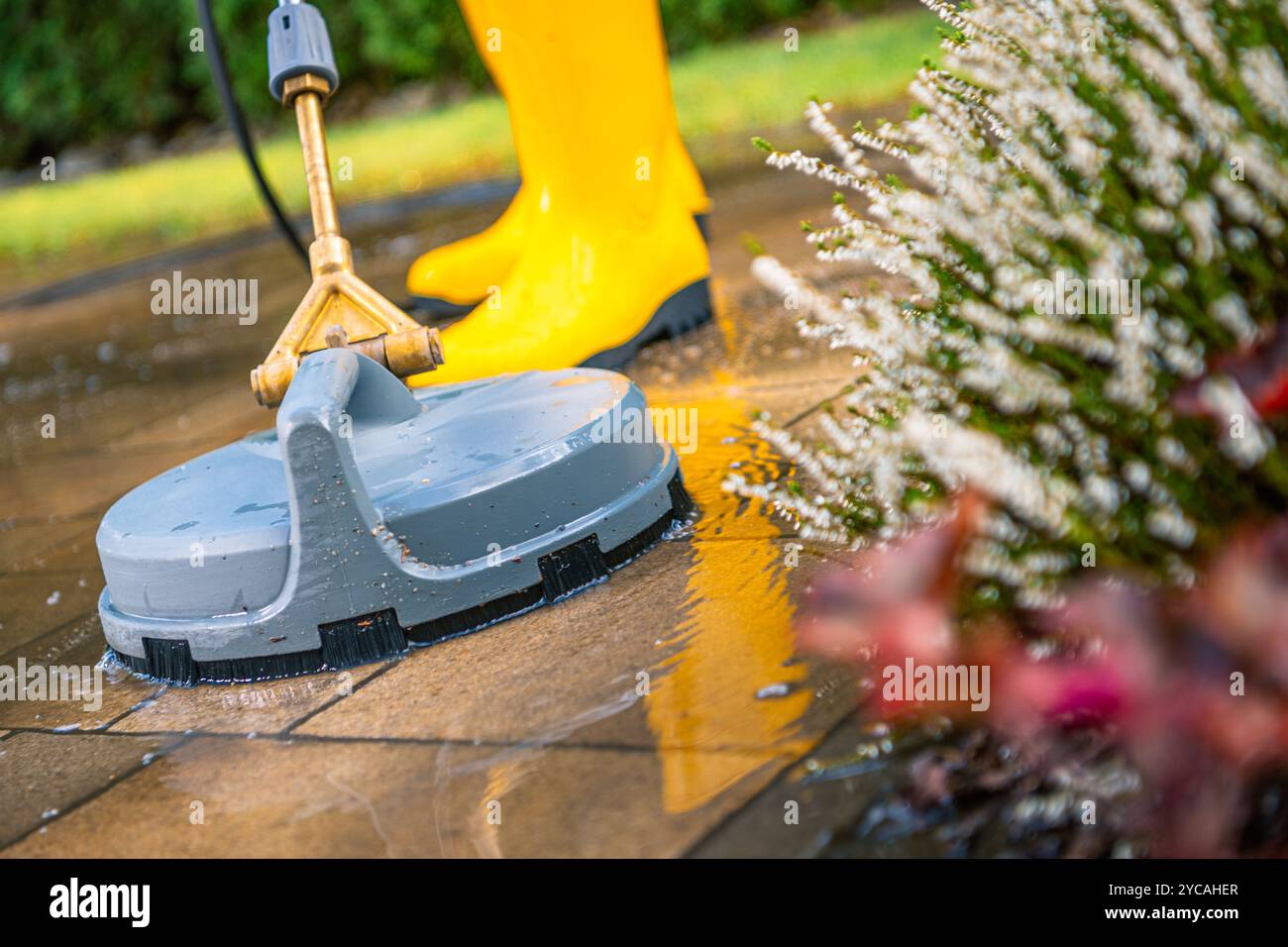 Una persona con stivali gialli utilizza una pulitrice a pressione per pulire le piastrelle sporche del patio in un vivace ambiente all'aperto in una giornata di sole. Foto Stock