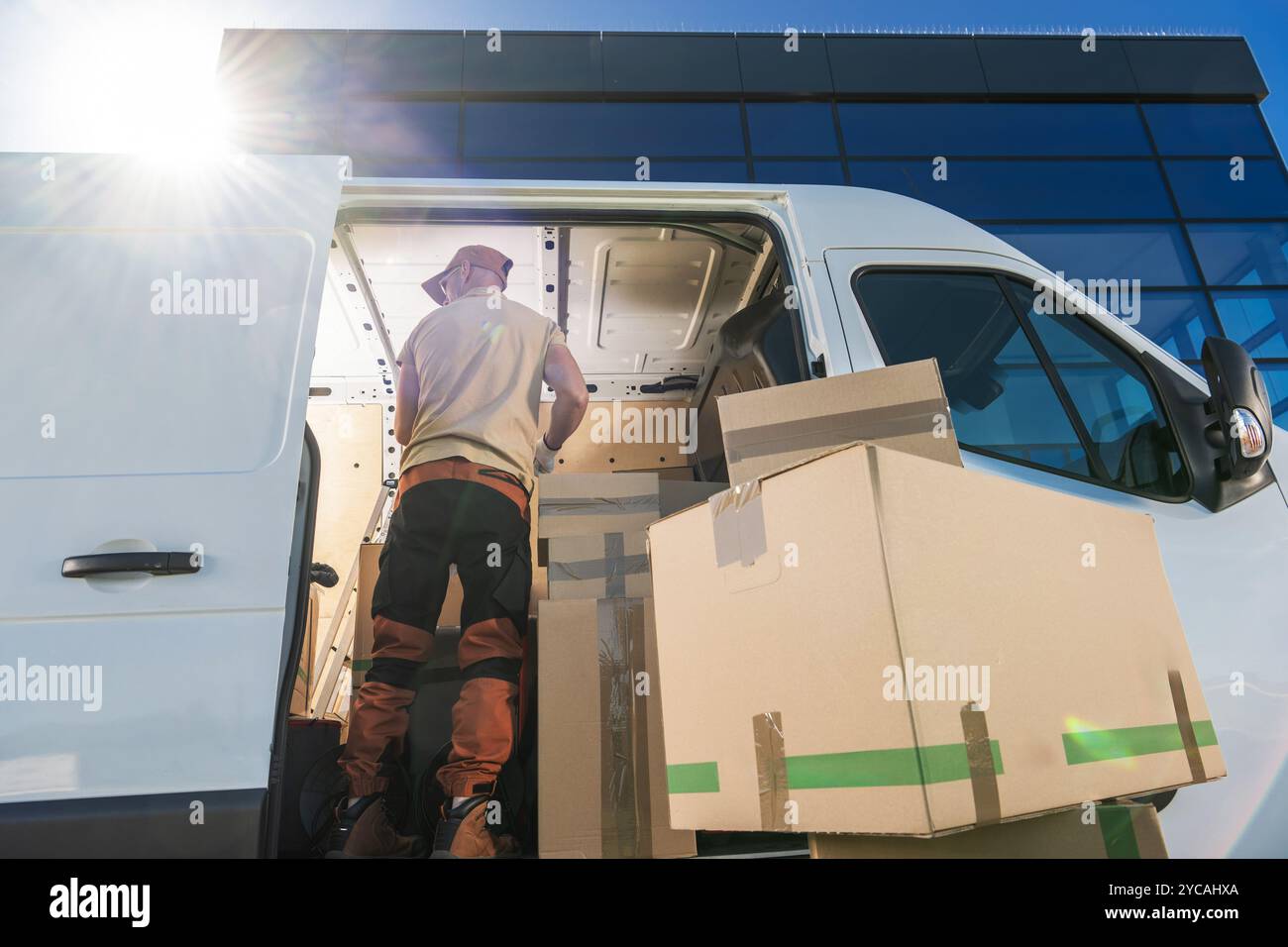 Un addetto alle consegne in abbigliamento casual scarica grandi scatole di cartone da un furgone parcheggiato in un ufficio contemporaneo. Il sole splende brillantemente, proiettando ombre. Foto Stock