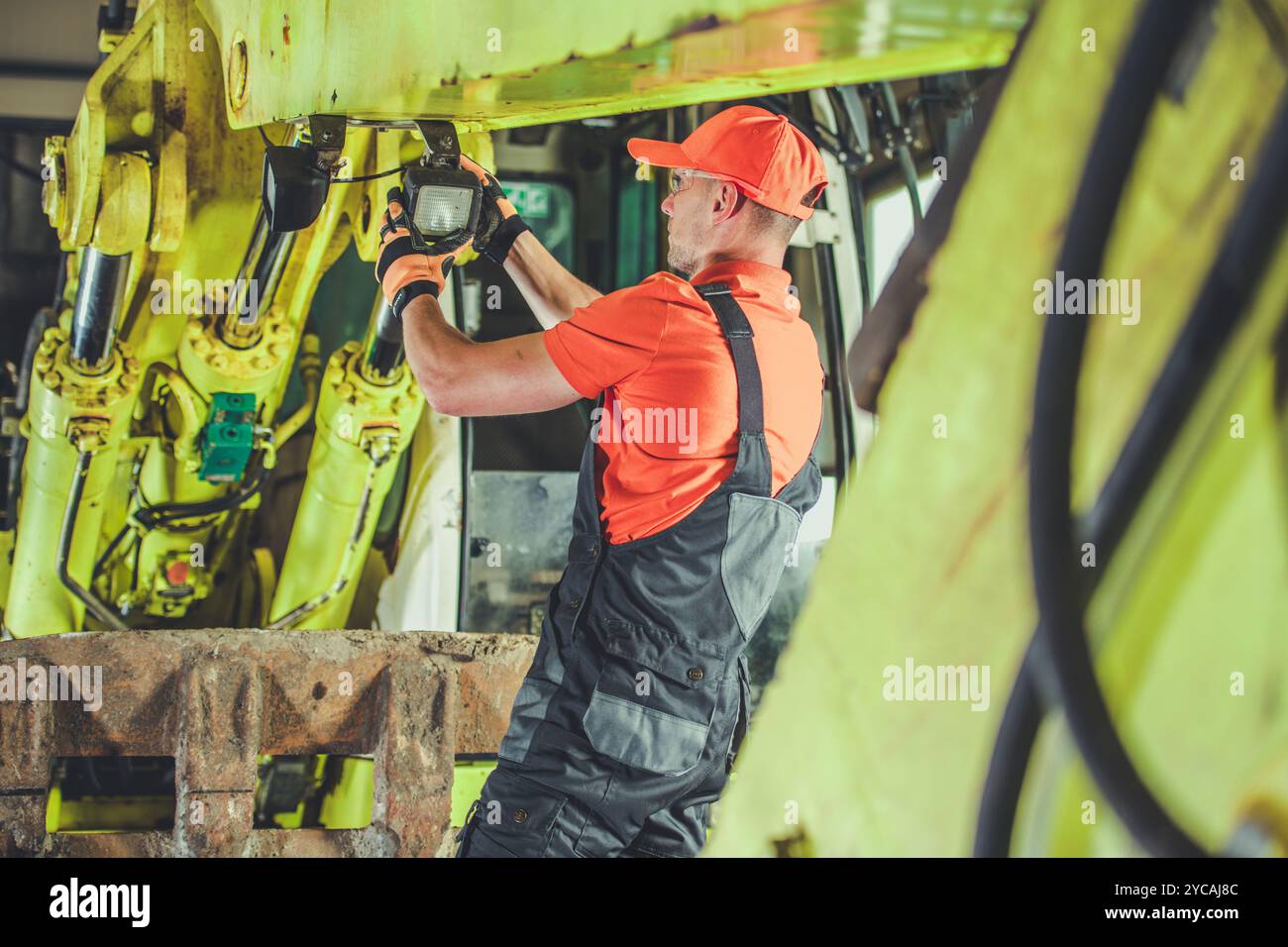 Un lavoratore vestito con una vivace attrezzatura di sicurezza ispeziona e ripara macchinari pesanti, garantendo il corretto funzionamento delle attrezzature. L'ambiente luminoso si evidenzia Foto Stock