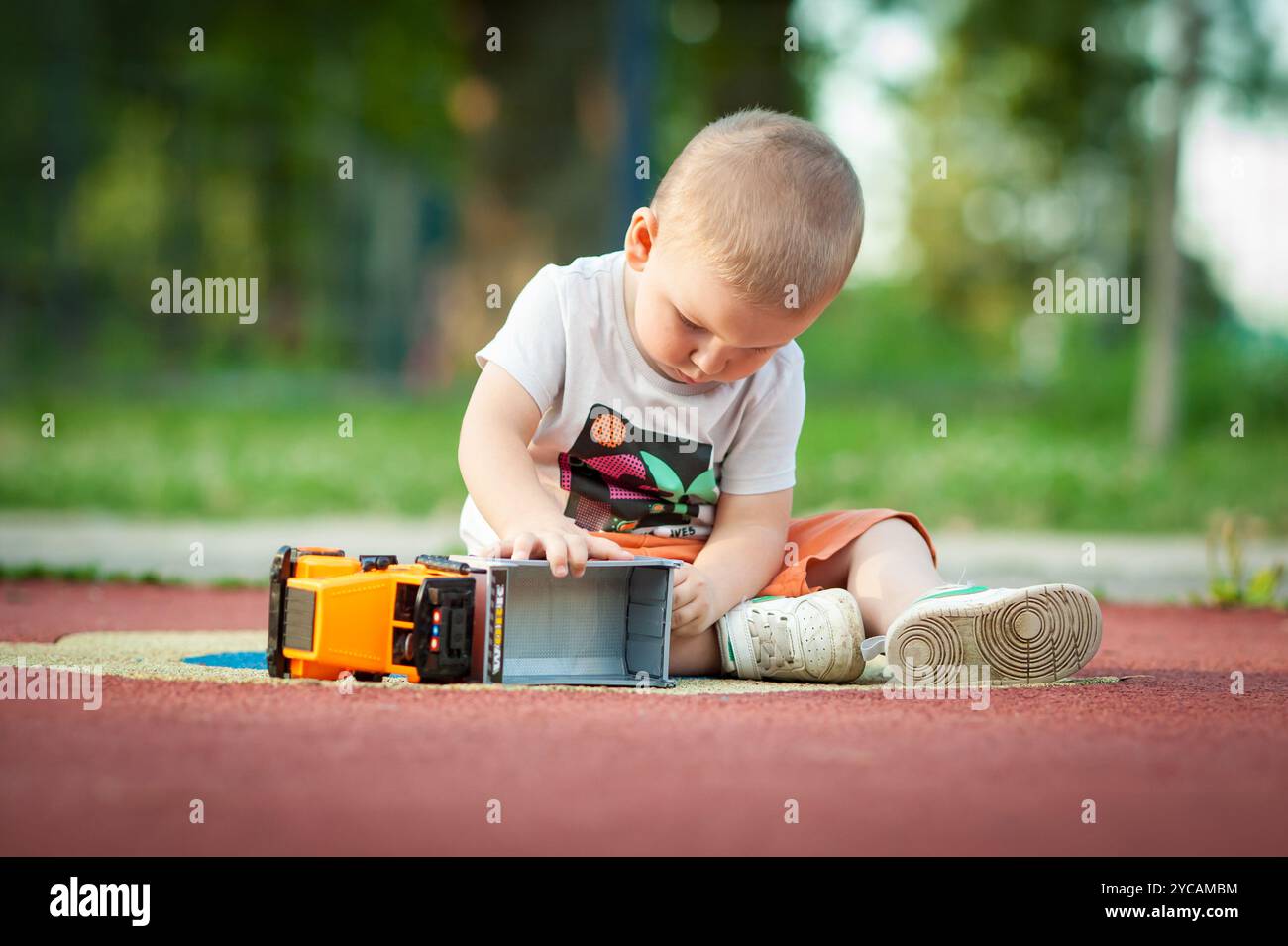 Un bambino si siede su un parco giochi colorato, incentrato sul gioco con un camion giocattolo. L'ambiente luminoso e la vegetazione esaltano la gioia Foto Stock