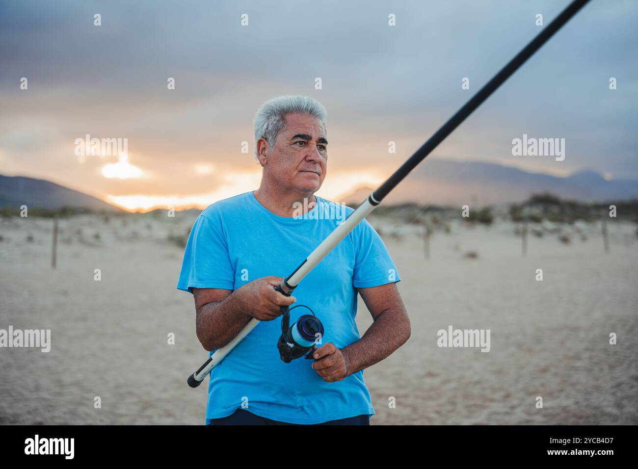 Un uomo maturo si trova su una spiaggia sabbiosa di Valencia, in Spagna, tenendo in mano una canna da pesca, con un tramonto sereno e montagne sullo sfondo Foto Stock