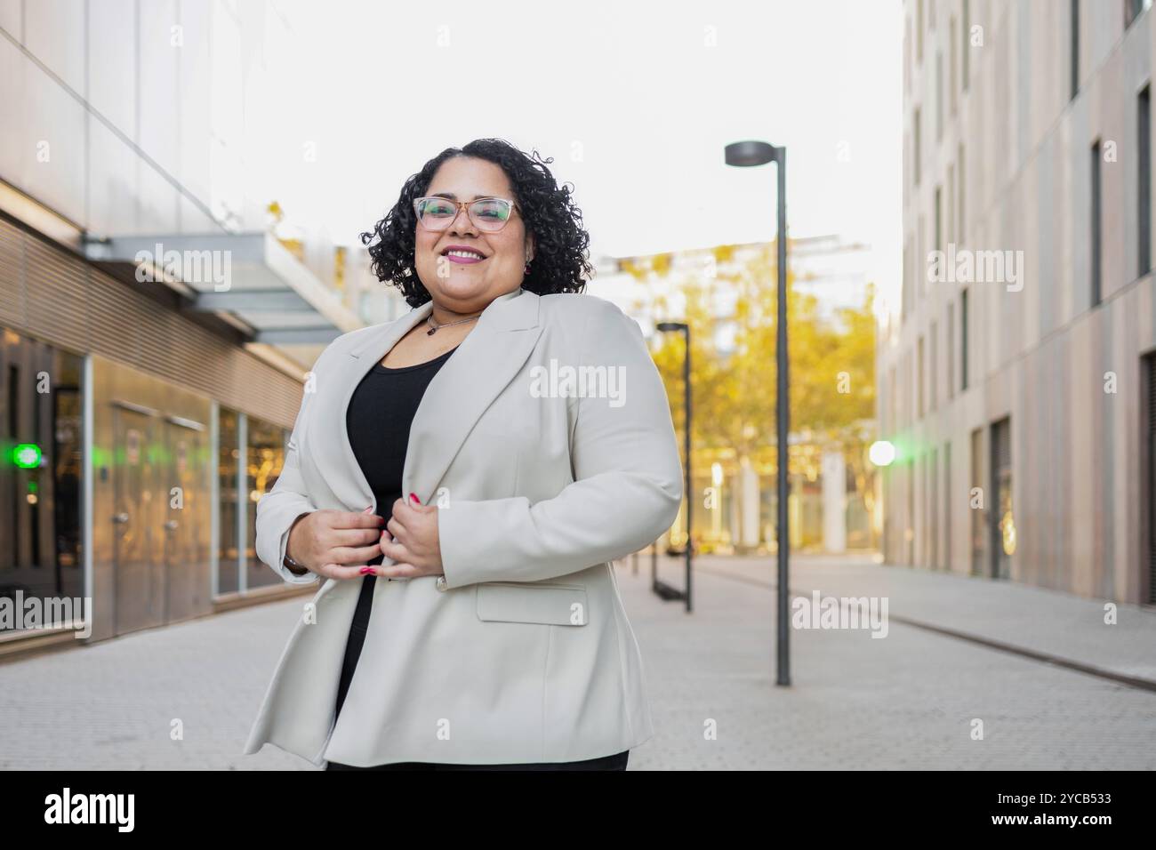Una donna d'affari ispanica con capelli ricci, occhiali e un blazer beige leggero sorride calorosamente alla fotocamera. Si trova con fiducia in un ur moderno Foto Stock