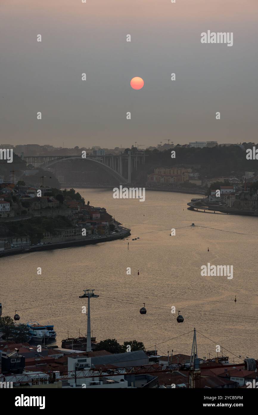 Immagine del sole che tramonta sull'iconico fiume Douro a Porto il tranquillo paesaggio è completato da sagome di funivie e dal ponte distante Foto Stock