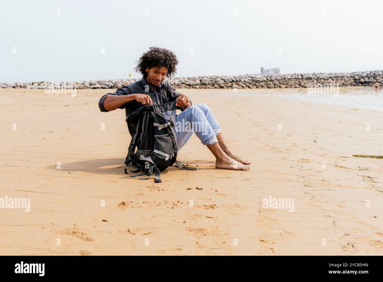 Un giovane con i capelli ricci si siede su una spiaggia sabbiosa, esaminando il contenuto del suo zaino sullo sfondo di mari calmi e di una lontana frangiflutti Foto Stock