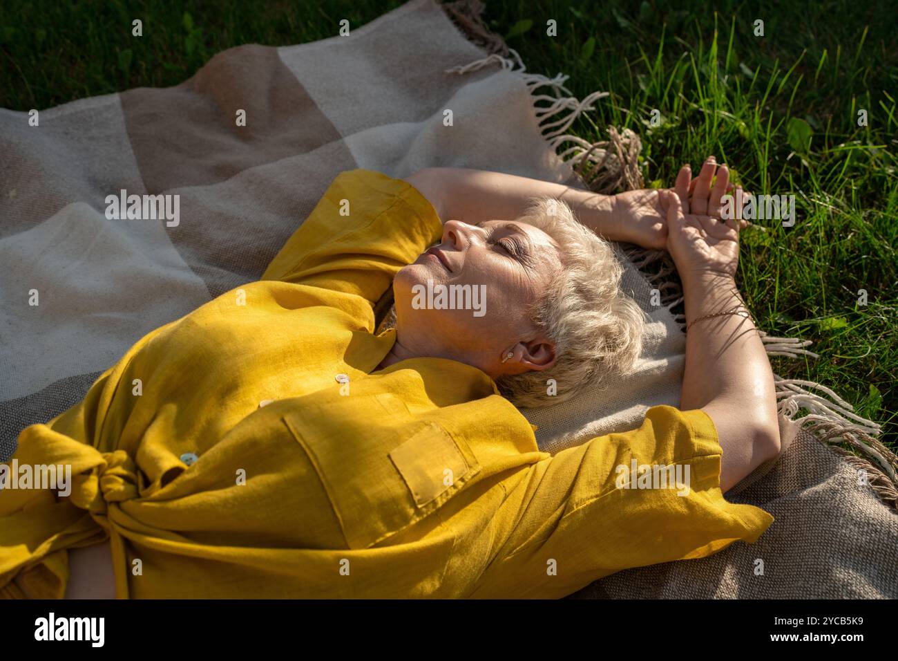 Una donna matura serena giace con gli occhi chiusi su una coperta in un campo, il sole che le illumina il viso, trasuda un'aura tranquilla in un ambiente naturale tranquillo. Foto Stock