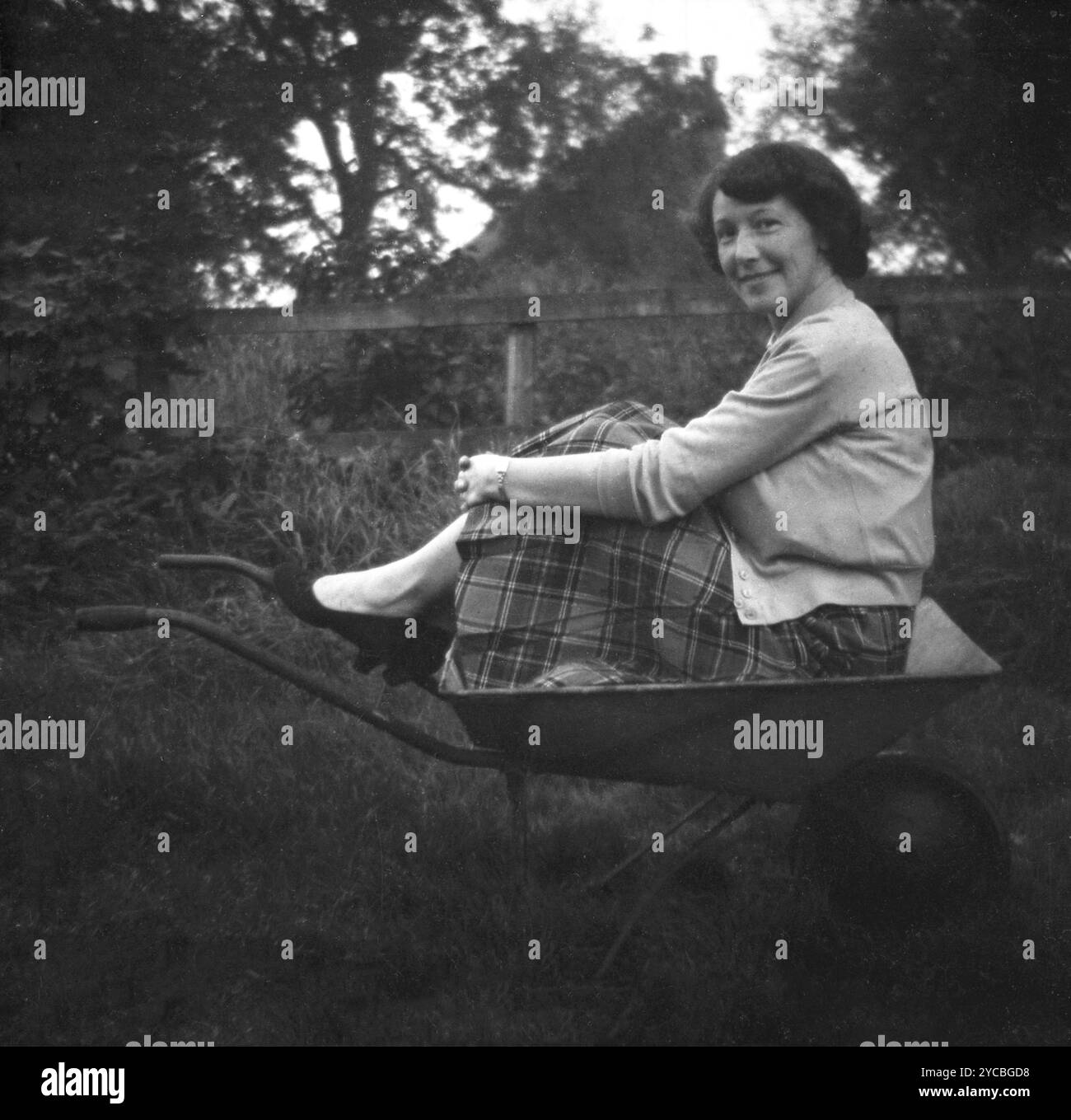 1950, storica, una giovane donna che indossa una gonna a pieghe tartan e un cardigan di lana seduto per una divertente foto all'aperto in una carriola da giardino... Foto Stock