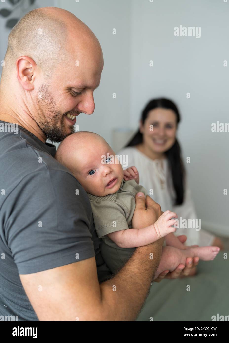 In primo piano sorride papà con neonato in braccio. Mamma si siede a letto e guarda le persone care. Al centro dell'attenzione c'è l'uomo. Famiglia felice. Foto Stock