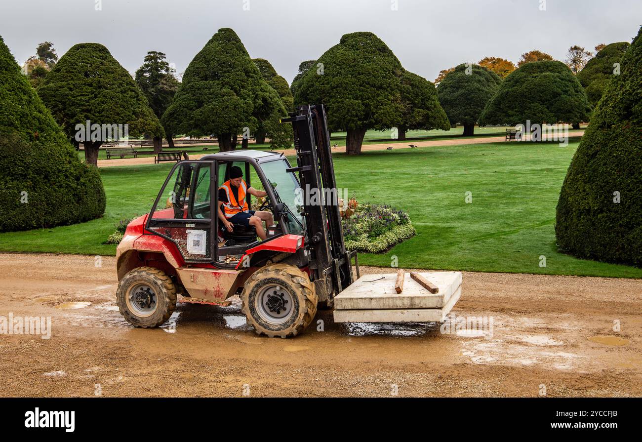 Un carrello elevatore con conducente in un giubbotto ad alta visibilità su un percorso sterrato circondato da siepi ben rifilate e un giardino ben tenuto Foto Stock