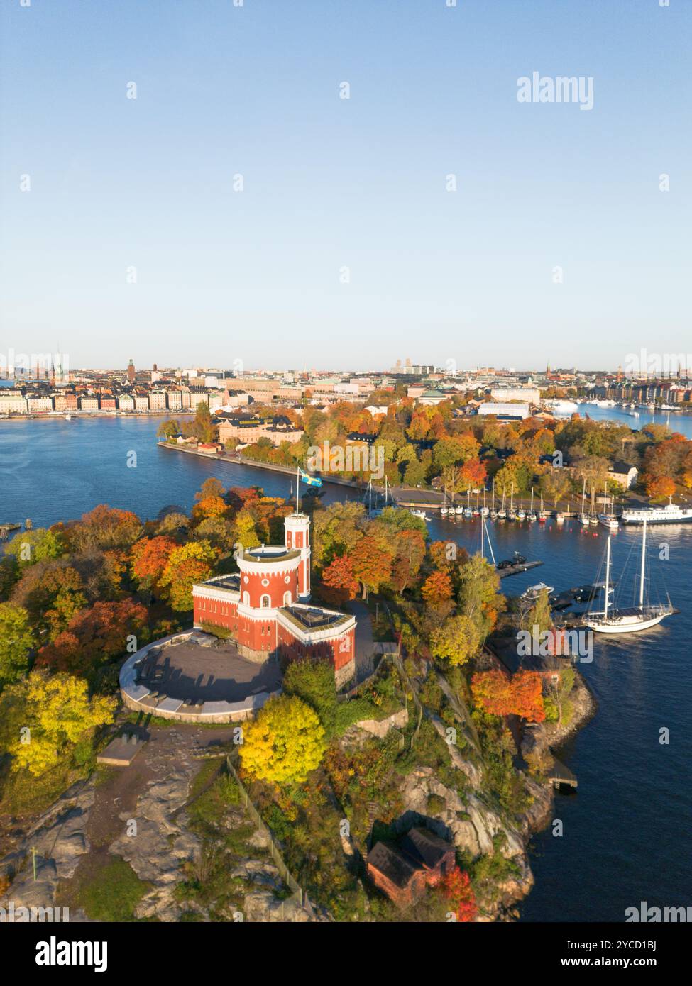 Castello rosso Kastellholmen sull'isola con lo skyline di Stoccolma sullo sfondo in una soleggiata mattinata autunnale con alberi gialli e aranci che si trasformano in Svezia Foto Stock