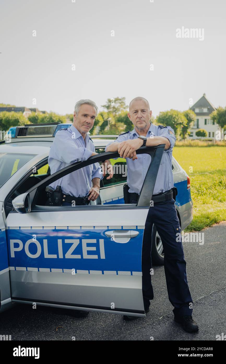 Colonia, Germania. 30 luglio 2024. Gli attori Heino Ferch (r) e Hannes Jaenicke stanno di fronte a un'auto della polizia durante le riprese. I due attori appaiono insieme davanti alla telecamera per la prima volta per la commedia ARD "Smeilingen". Credito: Marius Becker/dpa/Alamy Live News Foto Stock