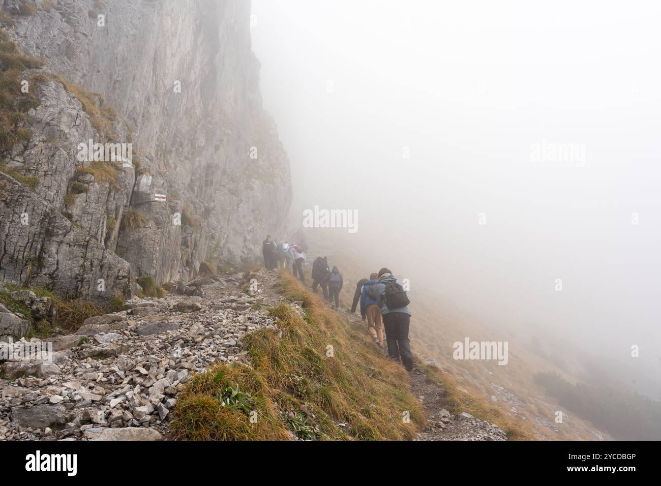 Gruppo di escursionisti che salgono uno stretto sentiero roccioso lungo un ripido pendio di montagna in una giornata nebbiosa. Concetto di avventura all'aperto, trekking in montagna e. Foto Stock