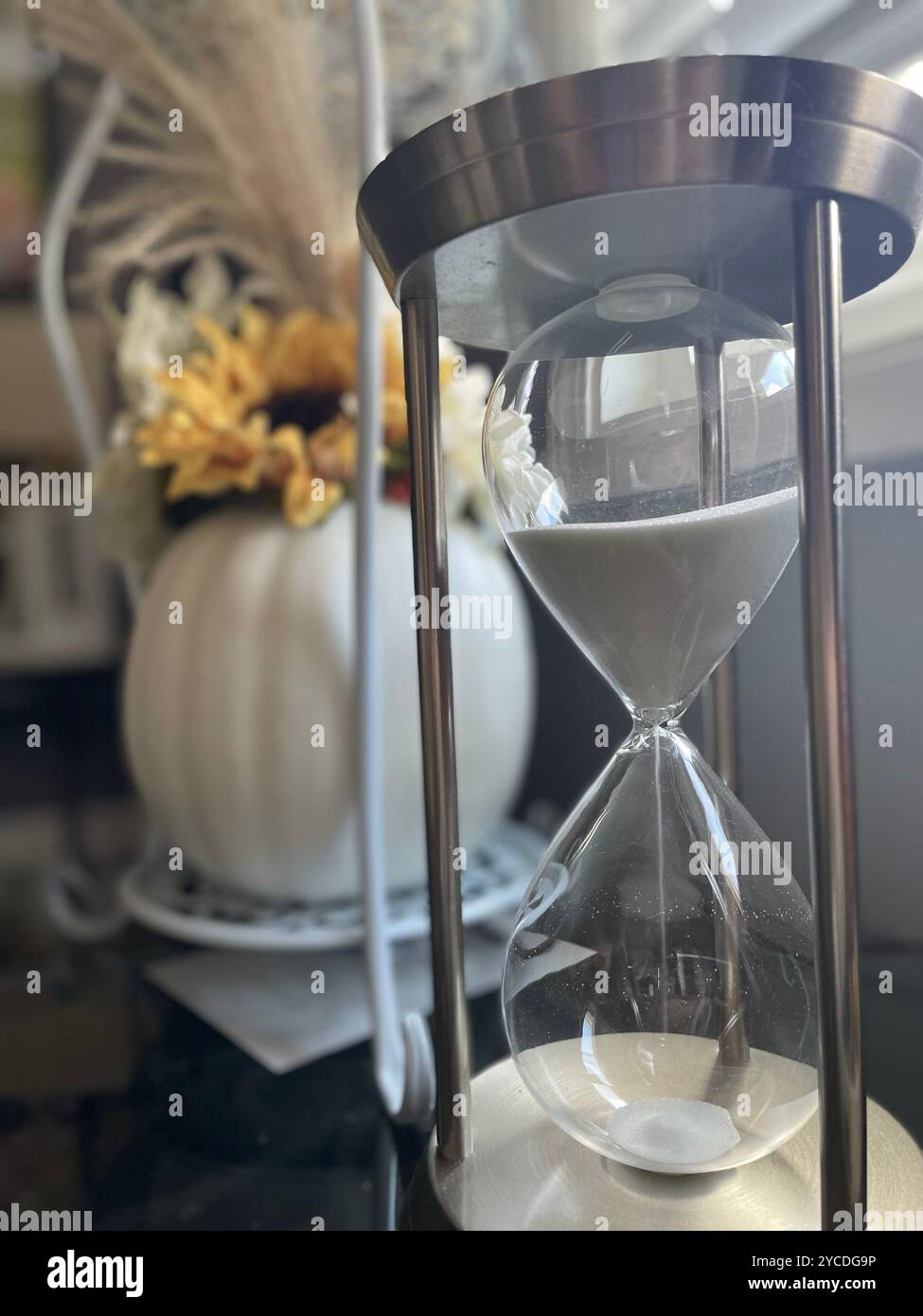L'elegante clessidra misura il tempo con la sabbia che scorre attraverso le lampadine di vetro. Sullo sfondo, un vaso sfocato con fiori gialli aggiunge calore alla scena. - Immagine stock catturata con smartphone