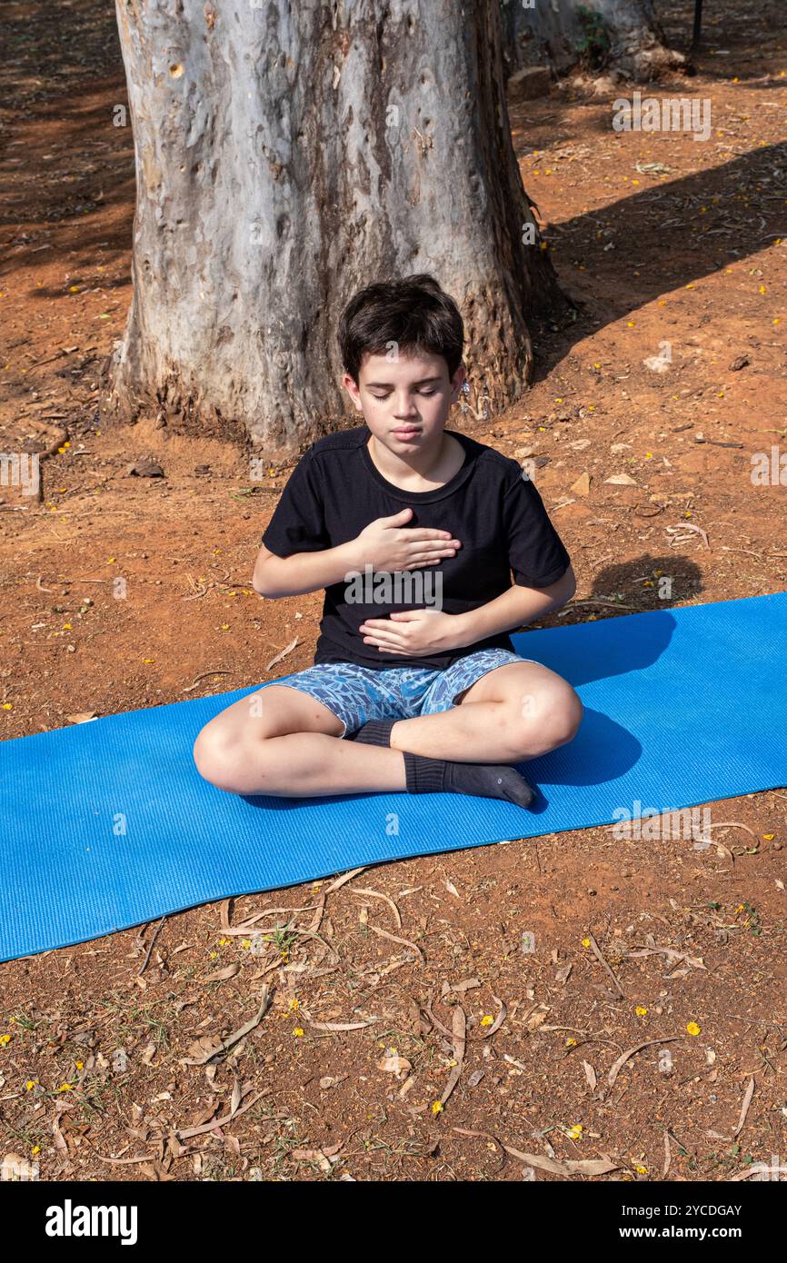 Bambino brasiliano seduto su un tappetino da yoga con le mani sul petto e l'addome nella classe all'aperto_5. Foto Stock