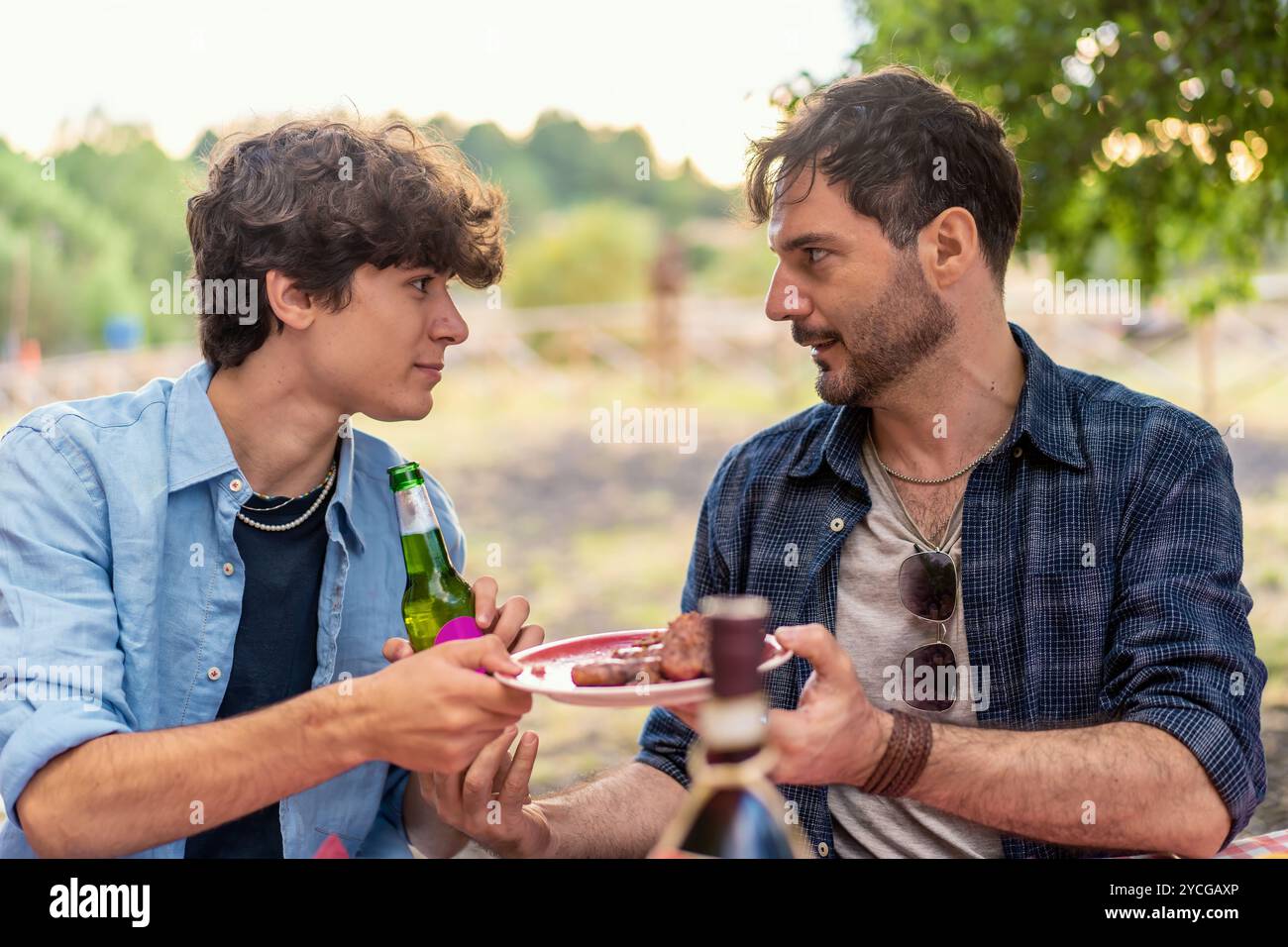 Padre e figlio adolescente hanno una conversazione significativa durante la riunione di famiglia all'aperto. Un naturale momento naturale di legame familiare con un pasto informale a nat Foto Stock