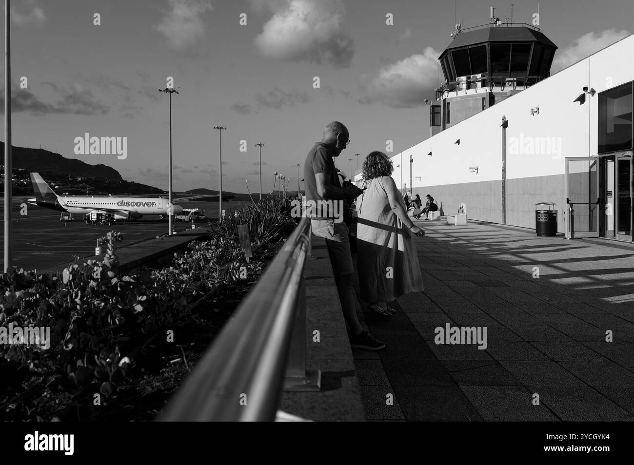 I passeggeri si rilassano presso la piattaforma di osservazione dell'aeroporto di Funchal, osservando gli aerei mentre il sole tramonta sull'isola in bianco e nero Foto Stock