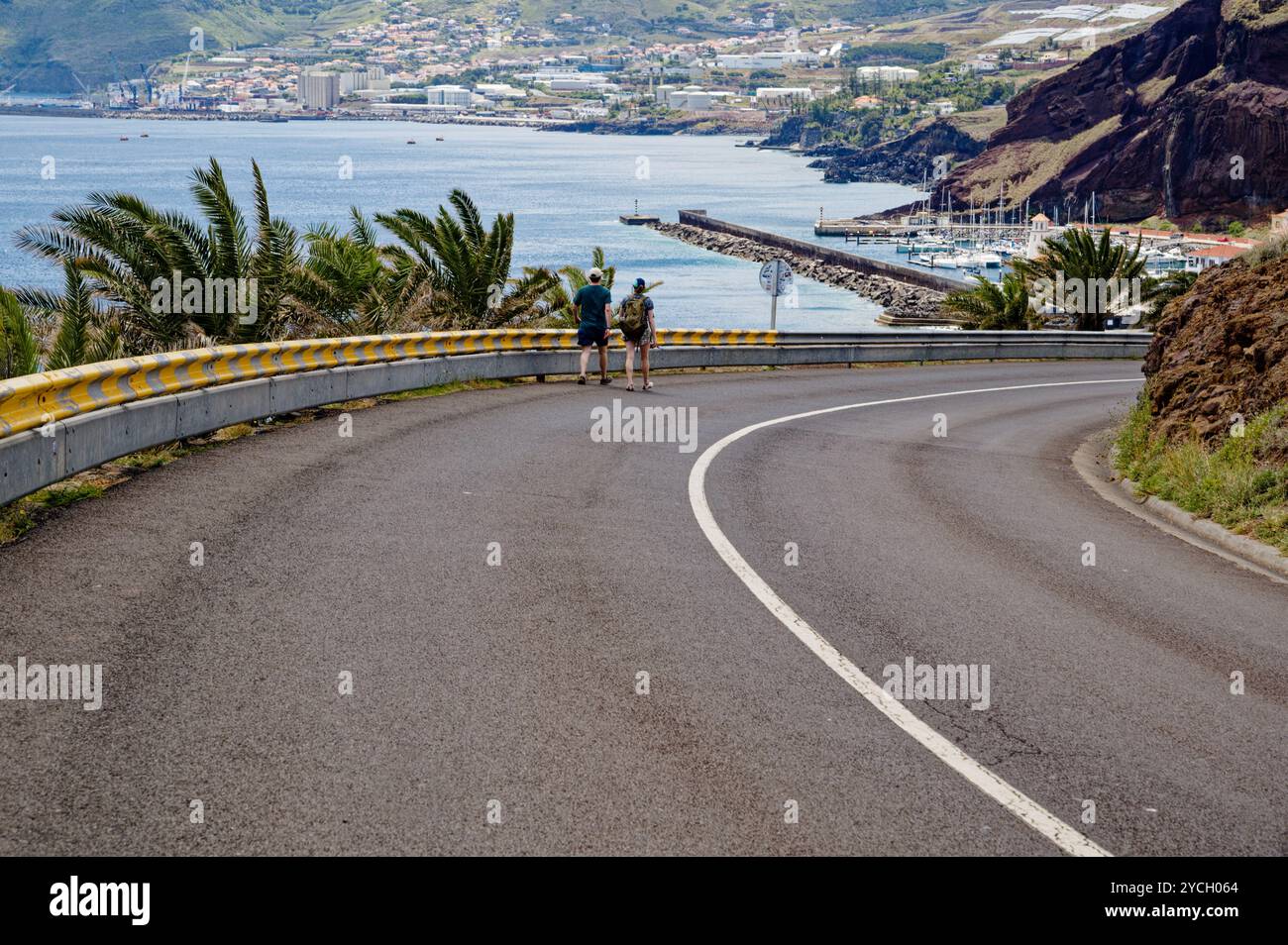 Due escursionisti che camminano lungo una strada curva che si affaccia sulla città costiera e sul porticciolo vicino a Vereda da Ponta de São Lourenco Foto Stock