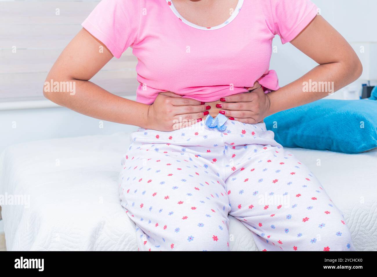 Primo piano di una giovane donna che preme l'addome mentre si siede su un letto, soffre di crampi mestruali. E' in una stanza ben illuminata Foto Stock