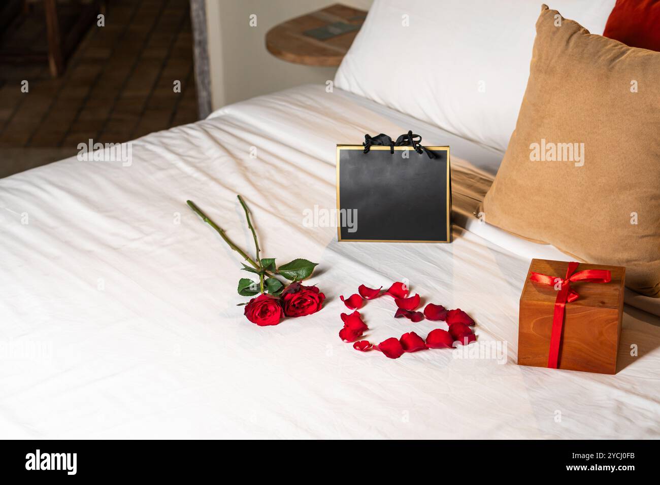 Un ambiente romantico con due rose rosse, un cuore a forma di petali di rosa, una scatola regalo con un nastro rosso e una borsa regalo nera su un copriletto bianco. Foto Stock