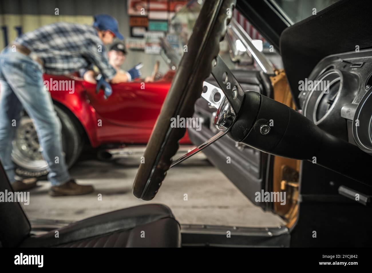 Un meccanico ripara con attenzione una classica auto sportiva rossa, mentre un'altra persona assiste nell'affollata officina automobilistica piena di attrezzi. Foto Stock