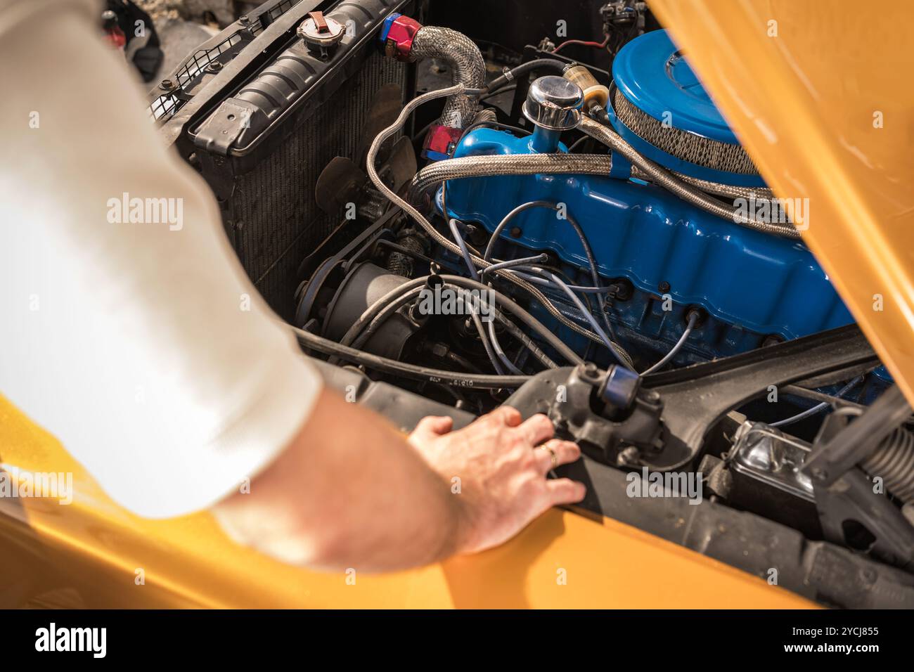 Una persona esamina il motore di un'auto d'epoca, concentrandosi su componenti dettagliati con attrezzi nelle vicinanze, mentre la luce del sole scorre attraverso il garage. Foto Stock