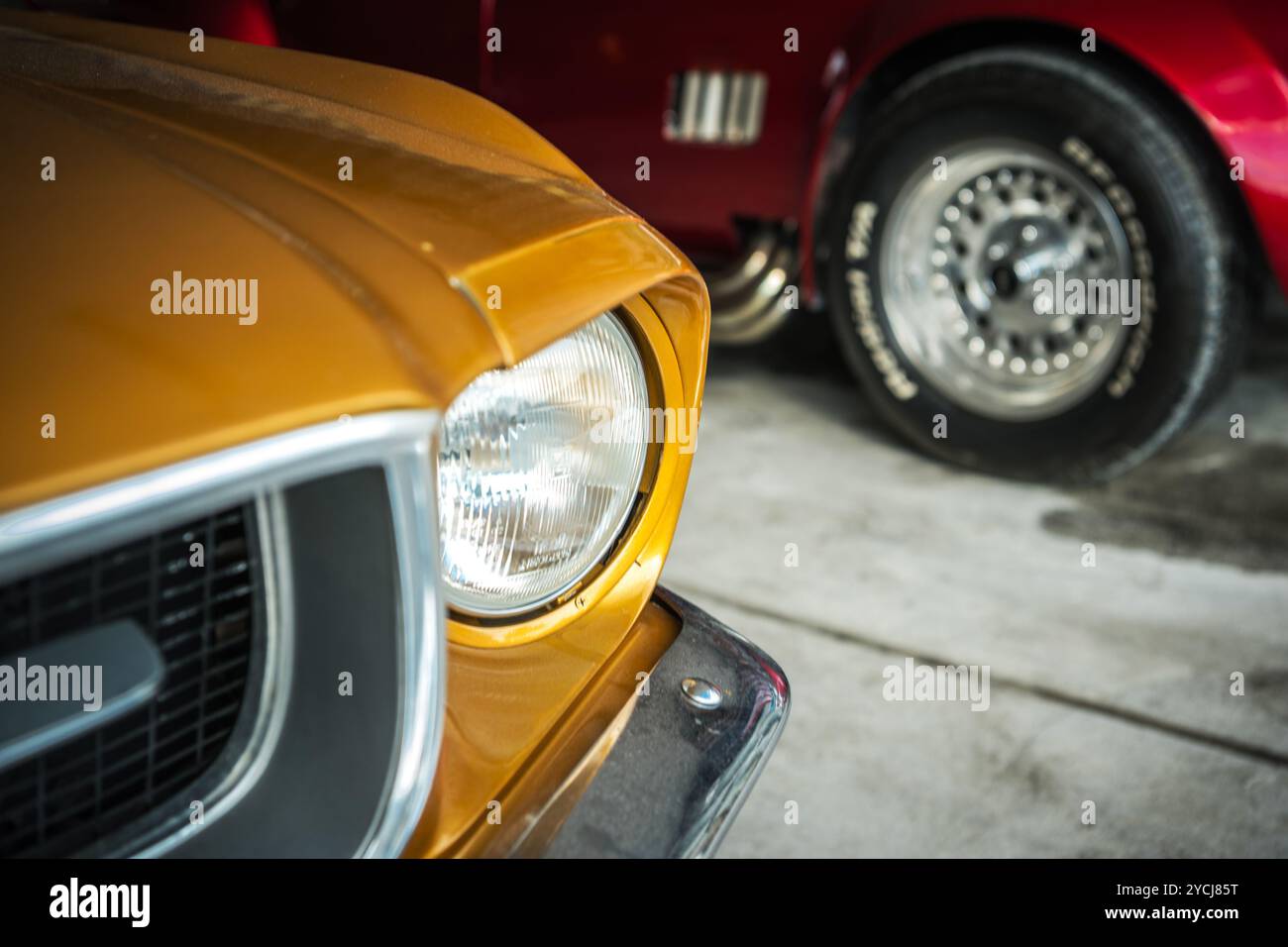 Vista ravvicinata della parte anteriore di una classica auto gialla accanto a un'auto rossa in un ampio garage, che enfatizza il design vintage e la maestria artigianale. Foto Stock
