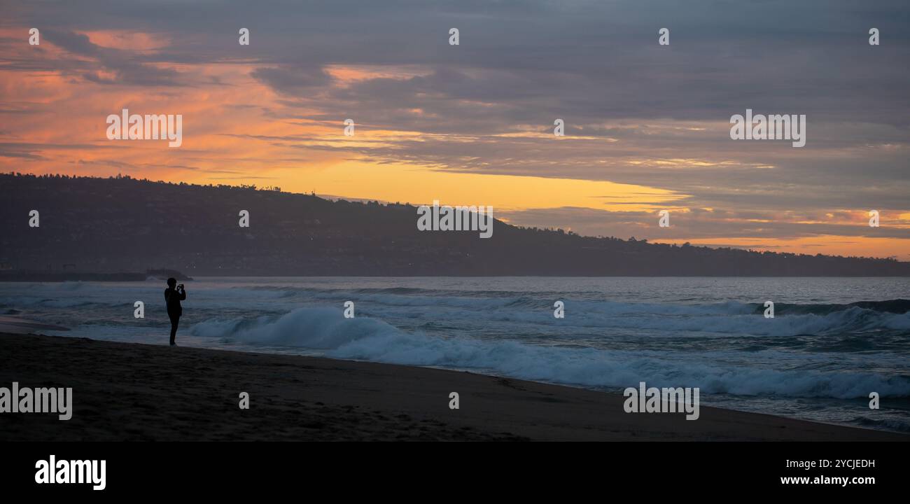 Persona con silhouette non identificabile che scatta foto al cellulare delle onde a Hermosa Beach, California, al tramonto con colline sullo sfondo Foto Stock