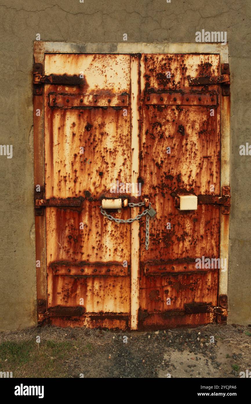 Una porta molto arrugginita chiusa a lucchetto su un edificio in cemento incrinato. Mi chiedo cosa ci sia dentro Foto Stock