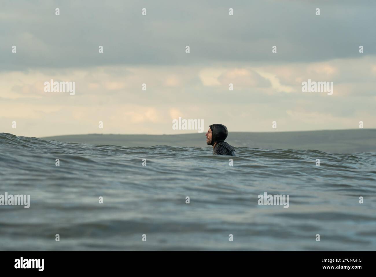 Un uomo con una muta nera con cappuccio siede su una tavola da surf in attesa di un'onda al largo di Bournemouth Beach, Bournemouth, Dorset, Regno Unito. Foto Stock