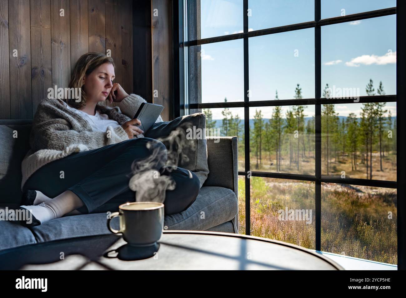 Donna seduta su un comodo divano, che legge un libro vicino a una grande finestra in una cabina con una bevanda calda accanto a lei, godendo la vista della foresta all'esterno Foto Stock