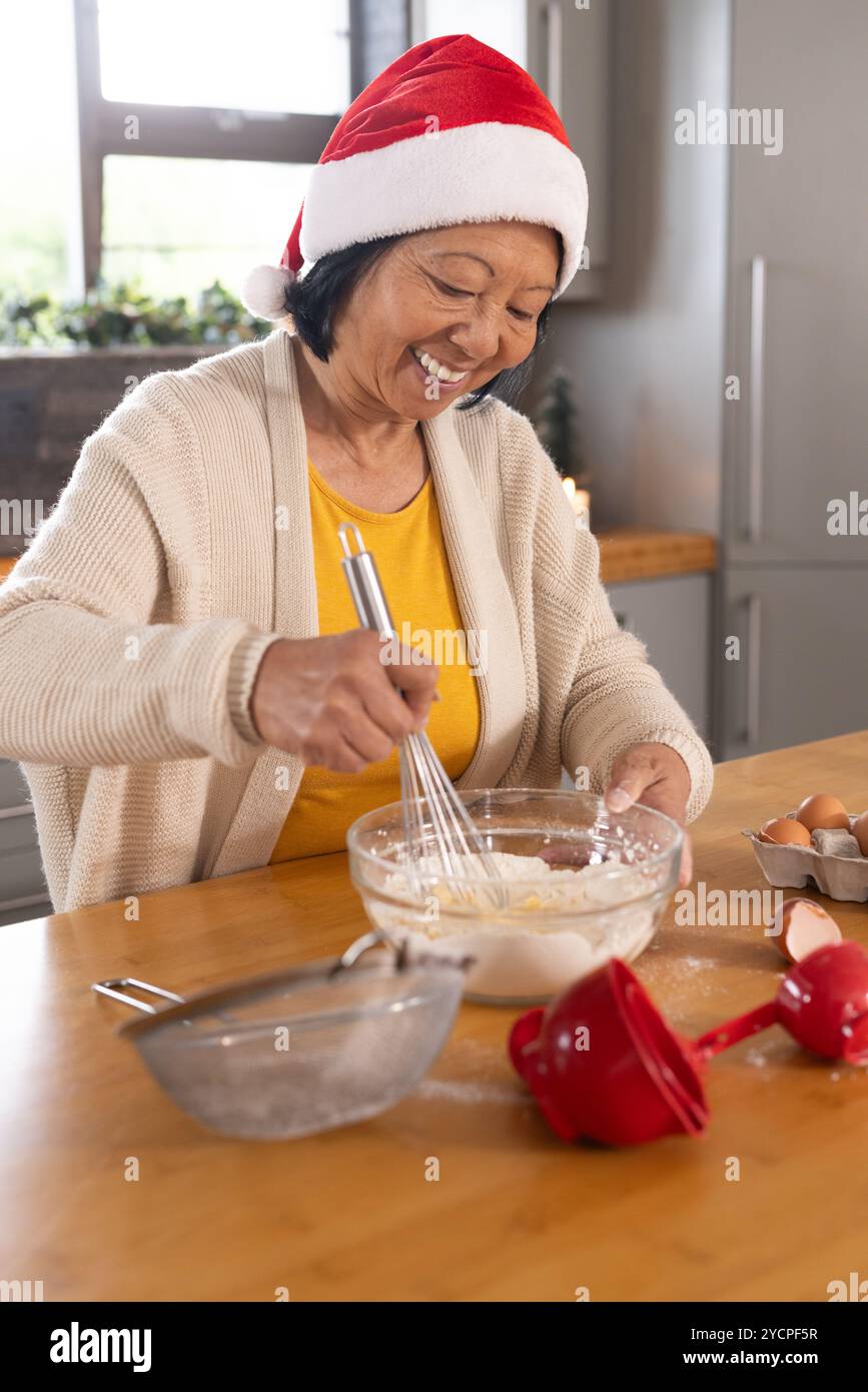 Donna asiatica anziana che indossa il cappello di Babbo Natale cuocere gioiosamente a casa durante il Natale Foto Stock