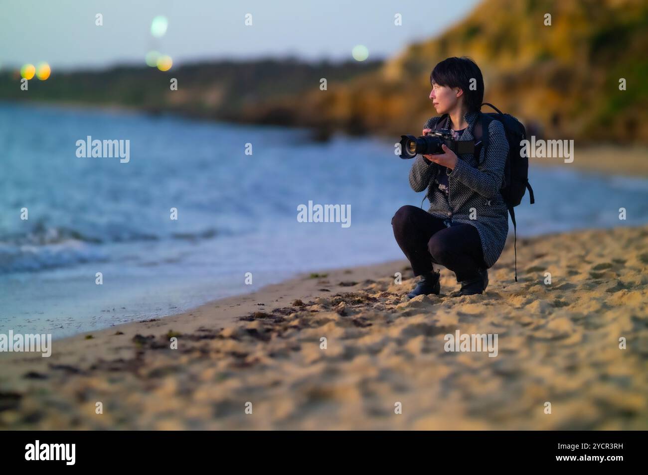 Una donna sta scattando delle foto a Sandringham Beach, Melbourne, Victoria, Australia, guardando lontano dalla macchina fotografica; luci della città sfocate in lontananza. Foto Stock